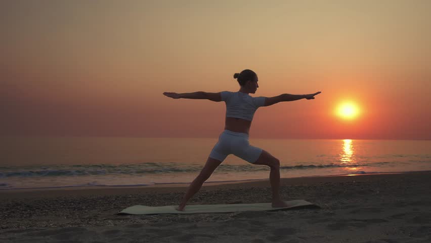 A woman practicing yoga in revolved triangle pose (parivrtta trikonasana) on a beach at sunset. Concept of balance, flexibility, mindfulness, and healthy lifestyle in a serene natural setting.
