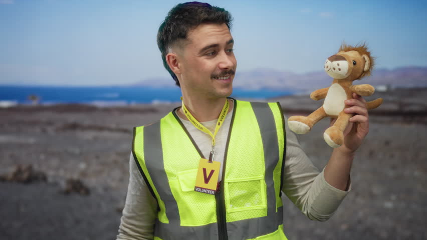 Young hispanic man wearing reflective vest holds plush toy on seaside beach showing volunteer badge with ocean view in background its blue sky emphasizing community effort