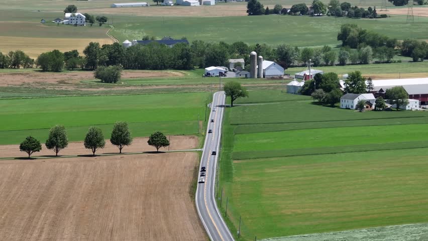 Traffic scene on rural highway of American town. Aerial zoom shot. Farmstead with houses and silo storage on homestead. Wide shot. Agricultural farm fields with forest trees. American countryside.
