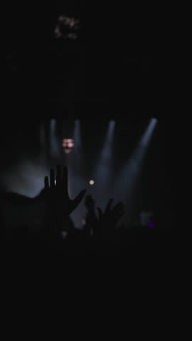 Strobe light show. Dark silhouette of raised hands of fans in wave motion. White, magenta and blue background light from stage. Medium close-up shot. Slow motion.