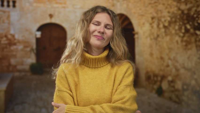 Woman holds hand while smiling in a courtyard portrait gesture with blonde talking wearing a yellow sweater against a stone wall.