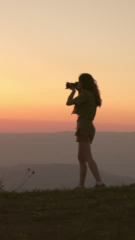 Female photographer standing on mountain summit at sunset, holding camera and capturing breathtaking landscape. Misty silhouettes of peaks create inspiring scene of freedom, creativity and slow travel