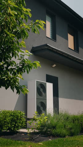 Vertical shot of an energy-efficient heat pump on a house wall, surrounded by greenery and shaded by tree branches in a landscaped garden. High quality 4k footage