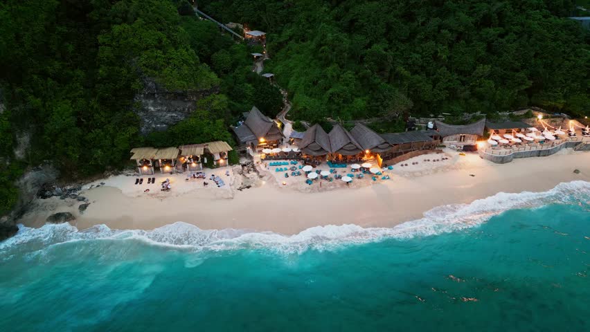 This drone video showcases a tropical dining spot by the sea with open-air seating on white sand, views of a turquoise ocean, and a towering cliff adding depth to the coastal beauty.