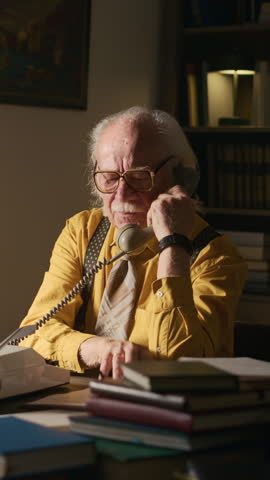 Vertical medium shot of grey haired Caucasian male writer in yellow shirt sitting at desk in front of typewriter and lots of books and dialing number on retro rotary phone calling publisher