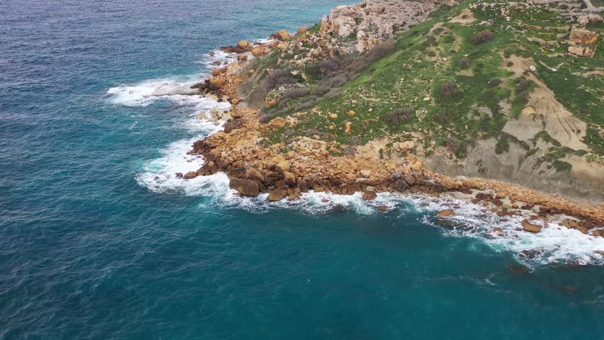 Aerial view of the rugged coastline meeting the turquoise sea, with golden rocks and green vegetation creating a vibrant contrast, Gozo Island, Gozo, Malta.