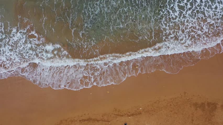 Aerial view of the sandy beach meeting the ocean, where foamy waves crash onto the shore in a mesmerizing dance of water and sand, Gozo Island, Gozo, Malta.