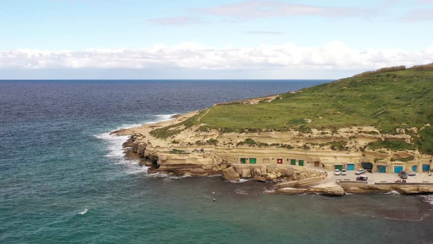 Aerial view of colorful boathouses nestled along a rugged coastline where the turquoise sea meets the golden cliffs topped with green vegetation, Gozo, Gozo, Malta.