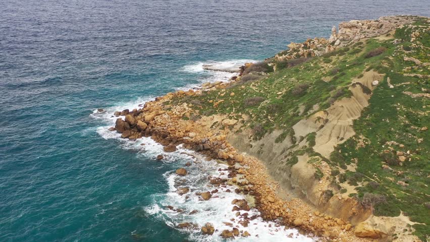 Aerial view of the rugged coastline where turquoise waters crash against the rocky cliffs covered with green vegetation, Gozo Island, Gozo, Malta.