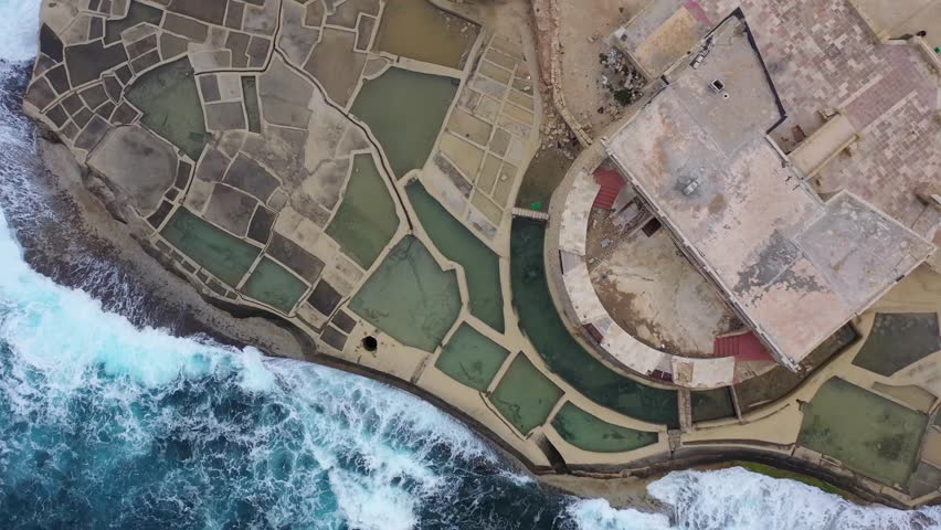 Aerial view of the geometric salt pans contrasting with the wild, churning turquoise waters crashing against the rocky shore, Gozo Island, Gozo, Malta.