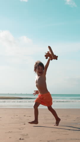 Happy little boy running along seaside with wooden toy airplane in hand, laughing and playing as if plane is flying. Slow motion scene of childhood joy, freedom, imagination and summer adventure on
