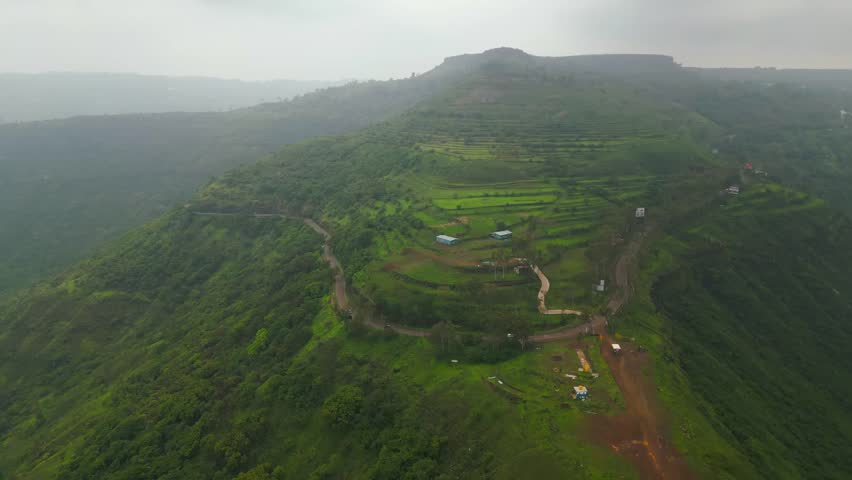 An aerial view of a ghat road in Panchgani, Maharashtra, shows vehicles driving through lush green mountains under a dramatic monsoon sky, capturing the vibrant aura of the season 4K