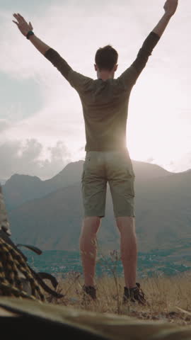 Male camper stretching arms joyfully outside tent at sunrise, facing majestic mountain landscape. Back view of traveler greeting morning with freedom, energy and healthy lifestyle during outdoor