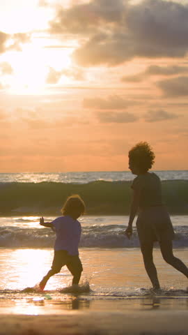 Mother and son playing tag on sandy beach at sunset, running joyfully along ocean shore. Slow motion scene of family love, freedom, childhood happiness and unforgettable summer memories in golden