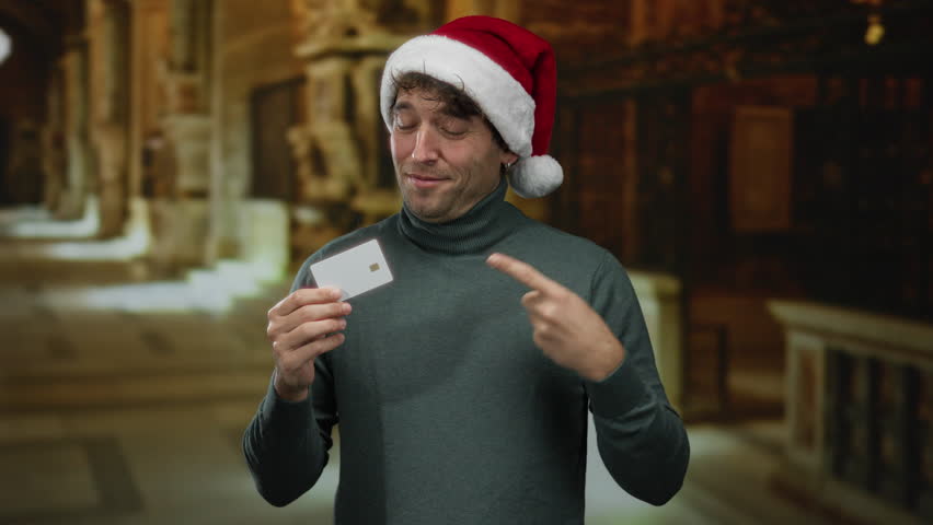 Man wearing christmas hat holds and points at credit card in museum gallery showing excitement and festive mood.
