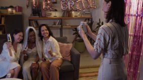 Young woman standing in front of her friends while taking photo via instant camera. Bride sitting on sofa surrounded by her bridesmaids, they smiling widely and holding champagne - Powered by Shutterstock - Get 15% off with code: PIKWIZARD15