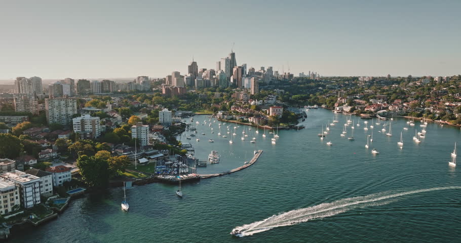 Australia, Sydney: Motorboat cruising Sydney Harbour, leaving white trail. Lavender Bay with sailboats yachts against the modern urban architecture city skyline in sunny day. Aerial view drone footage