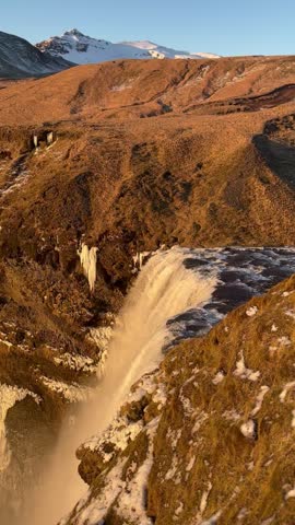 Aerial view of a majestic waterfall cascading down a rocky cliff in a snowy mountain landscape at sunset. Winter scenery with golden light and dramatic terrain.