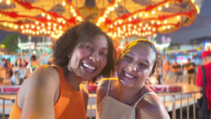 Two young women smile and pose for photos on a brightly lit carousel at a busy amusement park