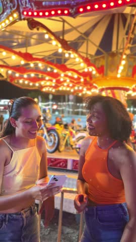 Two young women enjoy a fun moment at amusement park, sharing photos on a smartphone in front of a carousel