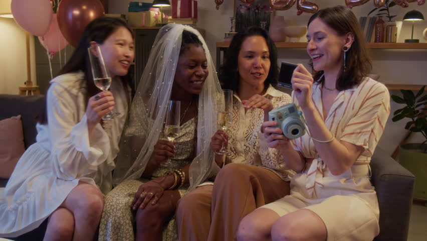 Bride with her best friends sitting together on sofa, holding glasses of champagne and looking instant photo