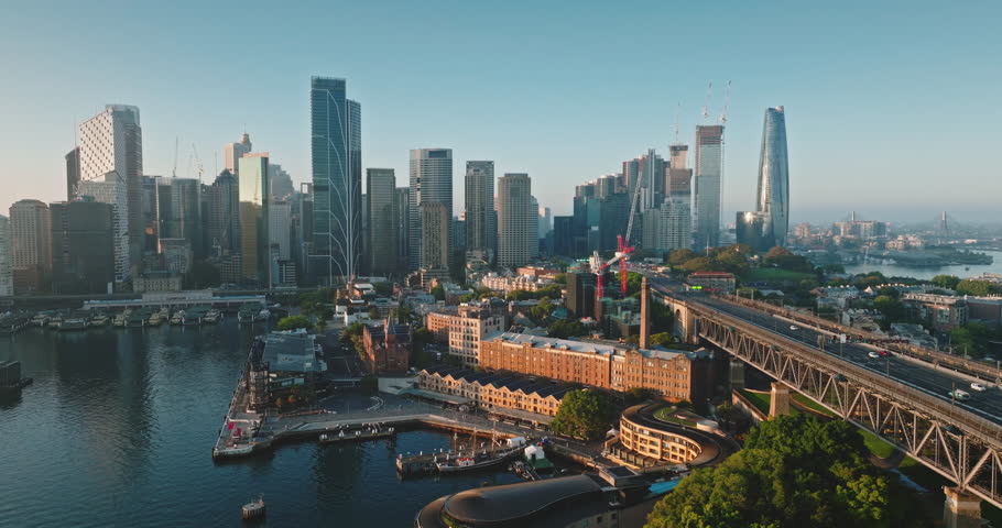 Australia, Sydney: Sydney Harbour Bridge connect business center city skyline with morning traffic, calm waters under blue sky. Modern architecture in background. Aerial view drone footage zoom out
