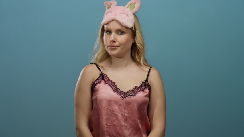 Young blonde woman in pink nightwear and bunny sleep mask gesturing while standing against a blue background, conveying a playful and watchful expression.