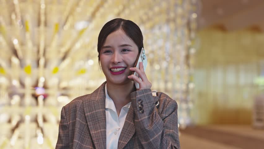 Asian Young woman smiling indoors, wearing plaid jacket and white shirt, communicating on phone with happiness and warm blurred background, gesturing with hand on chest
