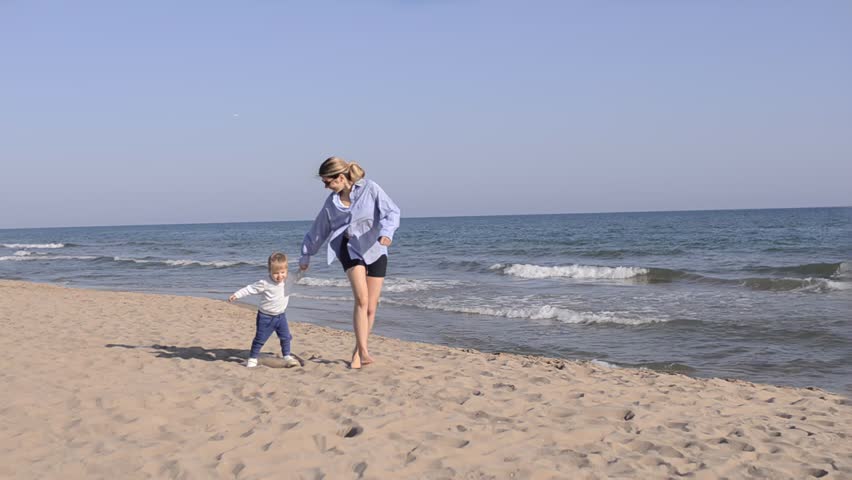 Happy mother and little boy running barefoot on sandy beach near sea	
