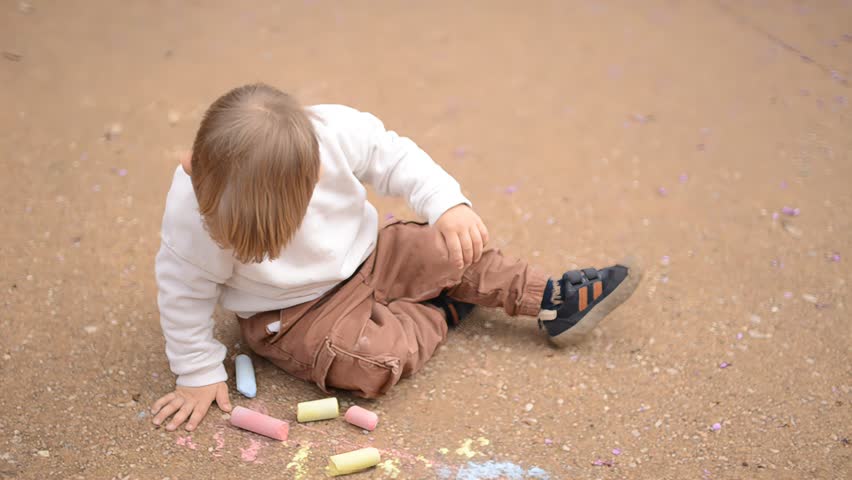 Young child holding chalk and sitting on sandy ground surrounded by colorful pieces in soft daylight	
