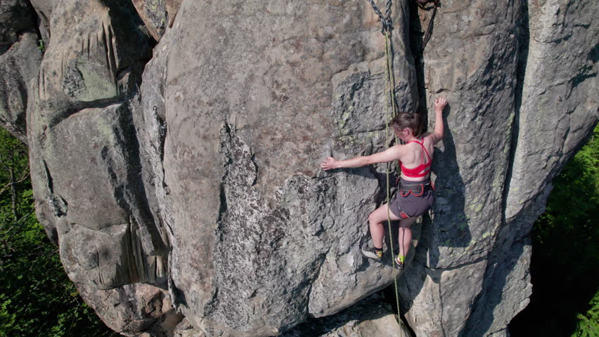Aerial view of woman rock climber ascending rugged limestone cliff with harness and rope for safety. Sportswoman climbing on vertical large boulder at Dobvush Rocks in Carpathian mountains, Ukraine.