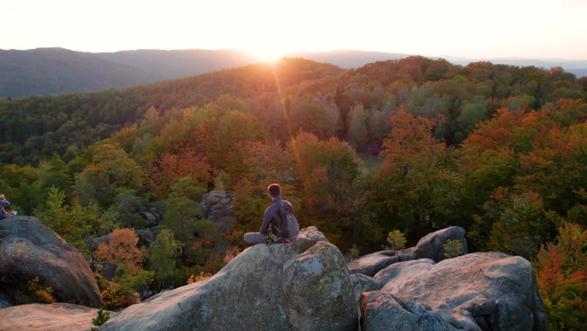 Aerial view of climber sitting atop rock formation at Dovbush Rocks in Carpathian mountains, Ukraine. Sun sets, casting warm glow over autumn forest and distant hills, creating breathtaking view.