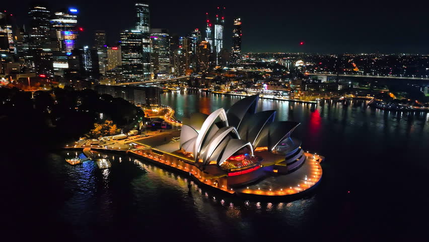 Australia, Sydney New South Wales: Sydney Opera House and city skyline at night, reflecting bright lights on water harbor surface, creating a breathtaking cityscape panorama. Aerial view drone footage