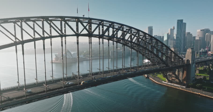 Australia, Sydney: Aerial view of Sydney Harbour Bridge, Sydney Opera House and city skyline in background. Sunny morning light, train and cars crossing the bridge. Modern architecture. Drone footage