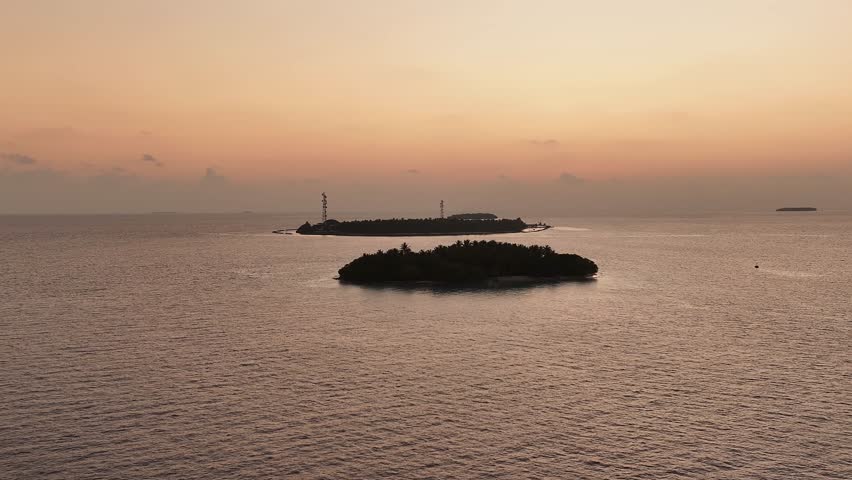 Aerial view of islands surrounded by calm waters under a pastel sunset sky casting a warm glow over the tranquil landscape, Kendhoo, Baa Atoll, Maldives.