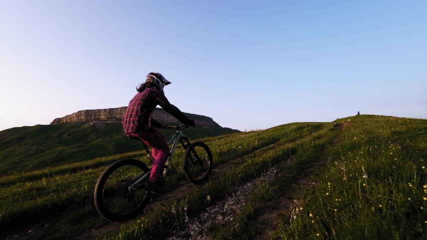 Young athlete cyclist in helmet and mask doing wheelie trick riding on rear wheel and pedaling in mountainous area at sunset. Aerial view shooting on fpv drone