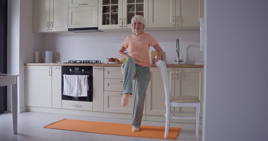 Happy sporty elderly woman with white hair doing stretching and morning exercises on rug in cozy kitchen at home. Sports activities and warm-up