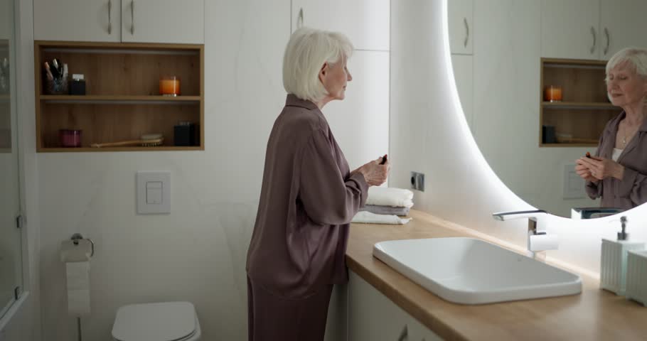 Zoom in elderly woman with white hair in purple pajamas doing makeup with lipstick in bathroom in the morning. Elderly woman doing make up in bathroom