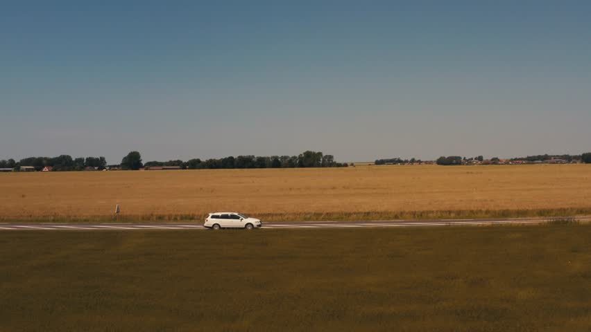 side view of car driving on road in middle of summer field, sun in background, blue sky, windmills. Estate car, white station wagon moving fast, aerial view filming camera drone. only one car alone.