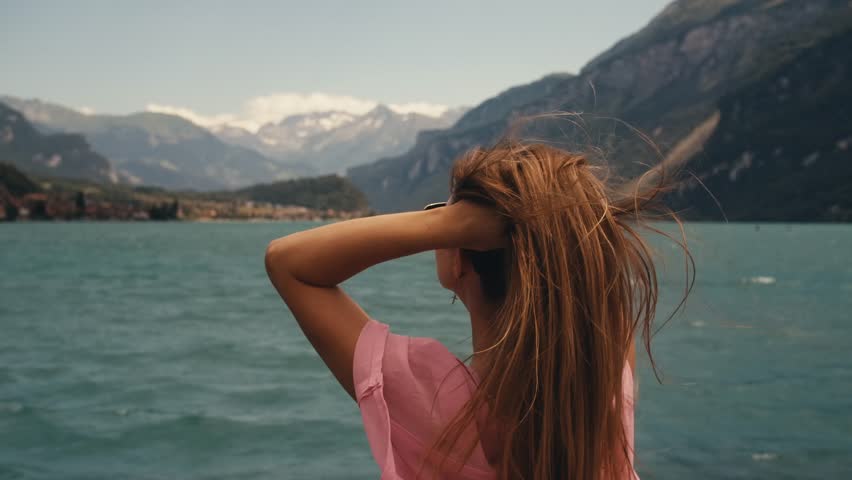 beautiful woman long blonde hair in white swimsuit pink pareo cape on standing on shore. summer background of blue sky lake mountains in Switzerland. slow motion wind blowing. Turquoise Azure Water