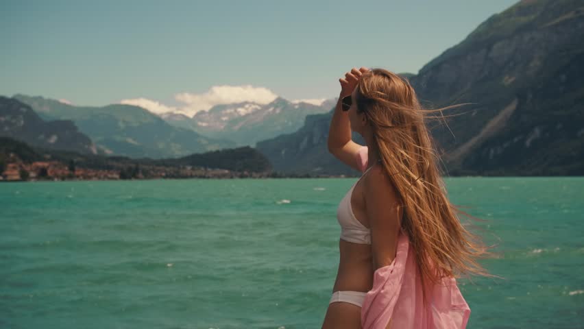 beautiful woman long blonde hair in white swimsuit pink pareo cape on standing on shore. summer background of blue sky lake mountains in Switzerland. slow motion wind blowing. Turquoise Azure Water