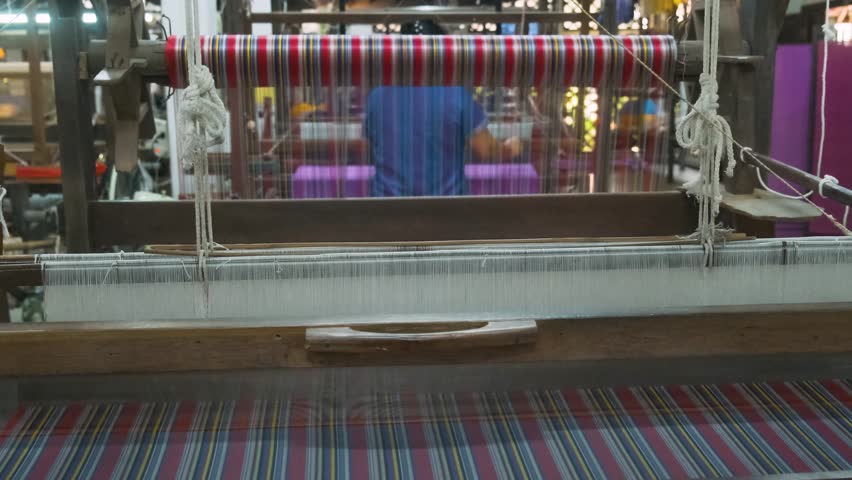 Colorful silk threads on loom with woman weaving in background on handloom in traditional silk factory, Chiang Mai, Thailand. Handmade craftsmanship, artistry of textile production