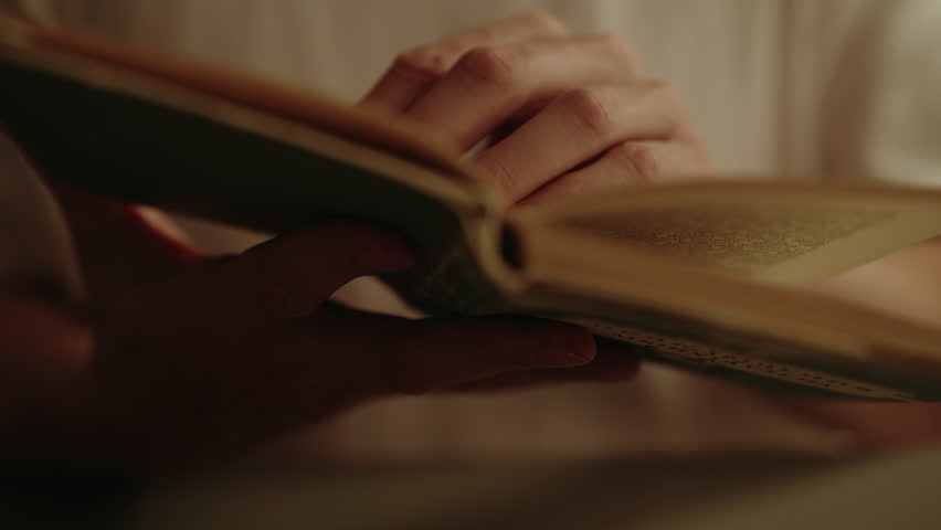 Close-up of young man hand turning page of old antique book in warm light at dark home setting, suggesting moment of peaceful immersion in literature. Concept of evening relaxation alone.