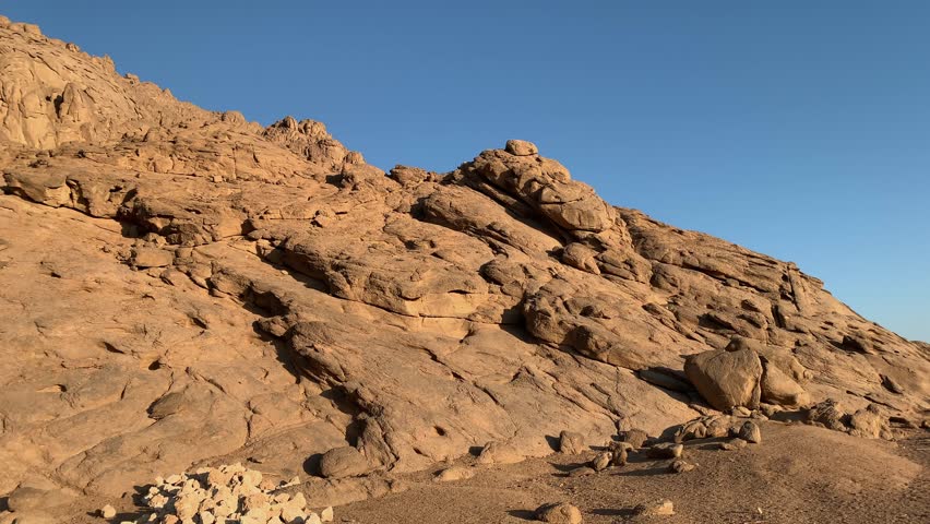 Arid desert terrain with large sandstone rock formations under warm sunset light. Rugged landscape with rocky hills and clear blue sky, evoking wilderness and remote natural beauty.