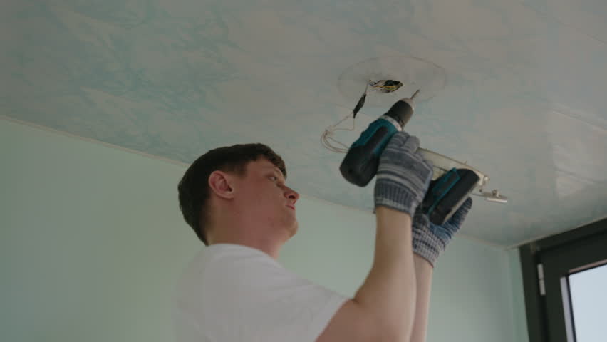 Skilled electrician male in protective gloves installing ceiling light fixture using power screwdriver ensuring secure electrical connections in room. Concept of working with lighting appliances.