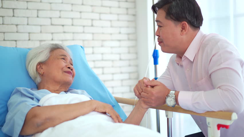 Asian man visiting elderly mother in hospital recovery room, holding hands and talking. Senior patient care, family support, modern healthcare concept.