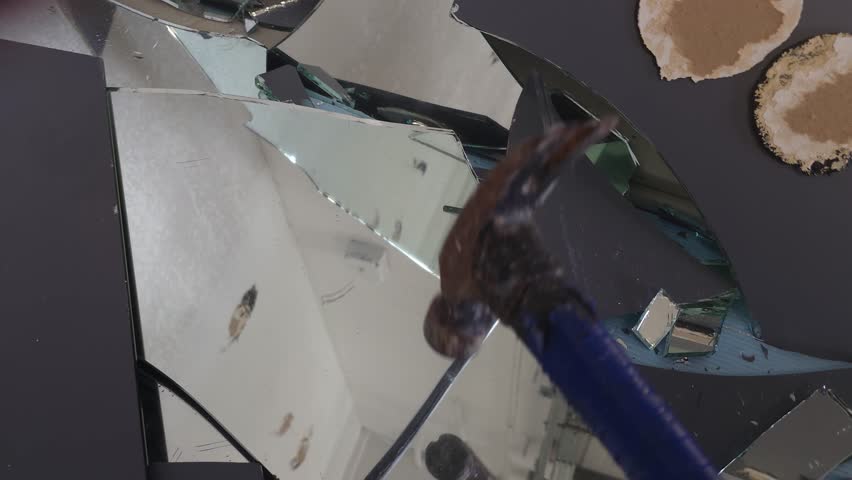 A hammer smashes cracked mirror shards lying on a blue surface. Fractured glass splinters scatter from the impact, highlighting destruction and danger.
