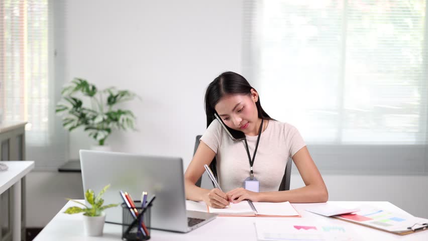 Professional businesswoman efficiently multitasking, writing notes while talking on phone, demonstrating workplace productivity and organizational skills at modern office desk - Powered by Shutterstock - Get 15% off with code: PIKWIZARD15