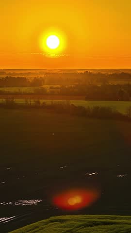 Bright golden sun rises above the horizon, casting a warm glow over vast, misty agricultural crop fields and rolling hills on a peaceful summer morning in Latvian countryside - revealing hyperlapse