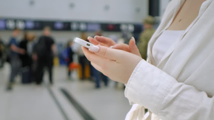 Woman is holding a cell phone in a busy airport. She is surrounded by people and luggage, and the atmosphere is bustling and hectic